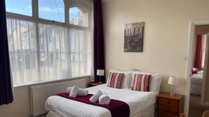 Un lit bien fait, avec du linge de lit blanc et des oreillers à rayures, une table de chevet avec une lampe, un radiateur et une fenêtre avec des rideaux.