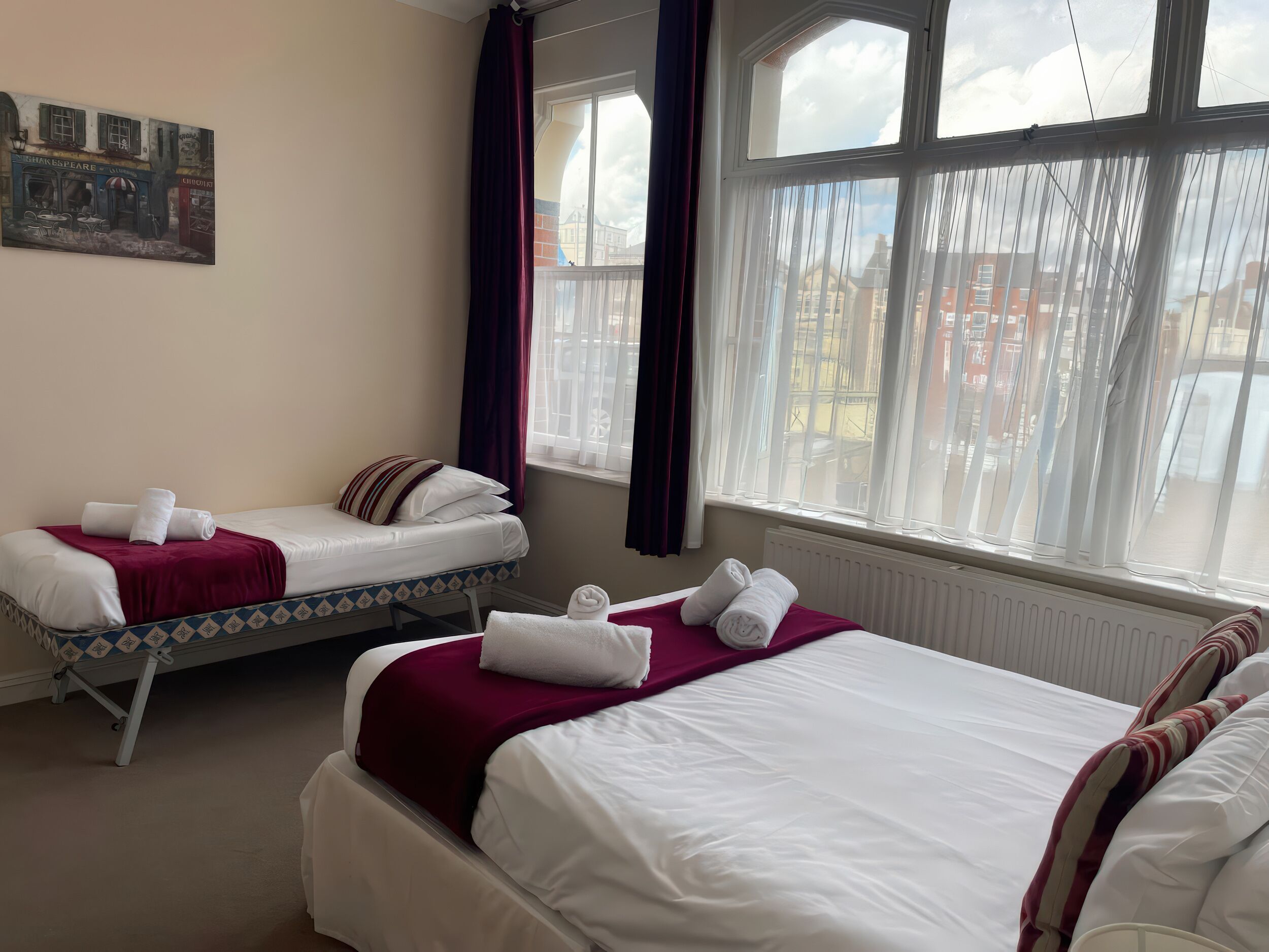 A hotel room with two beds, a large window with curtains, a painting on the wall, and a radiator.