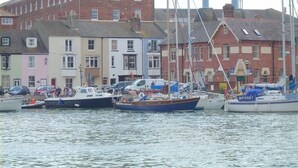 Un port de plaisance où sont amarrés plusieurs bateaux, un bâtiment en briques rouges et, au fond, des immeubles résidentiels aux couleurs vives.
