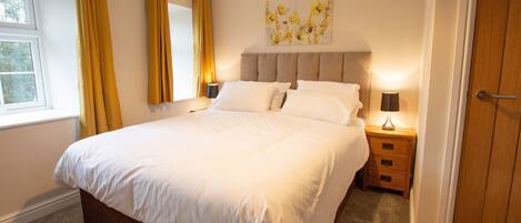 A neatly made bed in a hotel room with a window, a bedside table with a lamp, and a door.