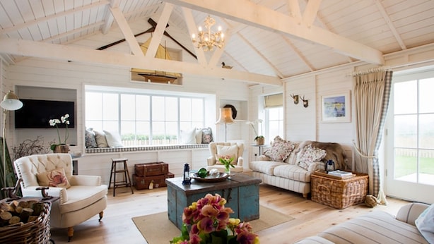A spacious living room with a high ceiling, wooden beams, a large window, a fireplace, and a television. - Lligwy Beach Cottage (Dulas)