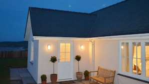 A white house with a dark roof, a front door, two potted plants, a bench, and a lit window. - Lligwy Beach Cottage (Dulas)