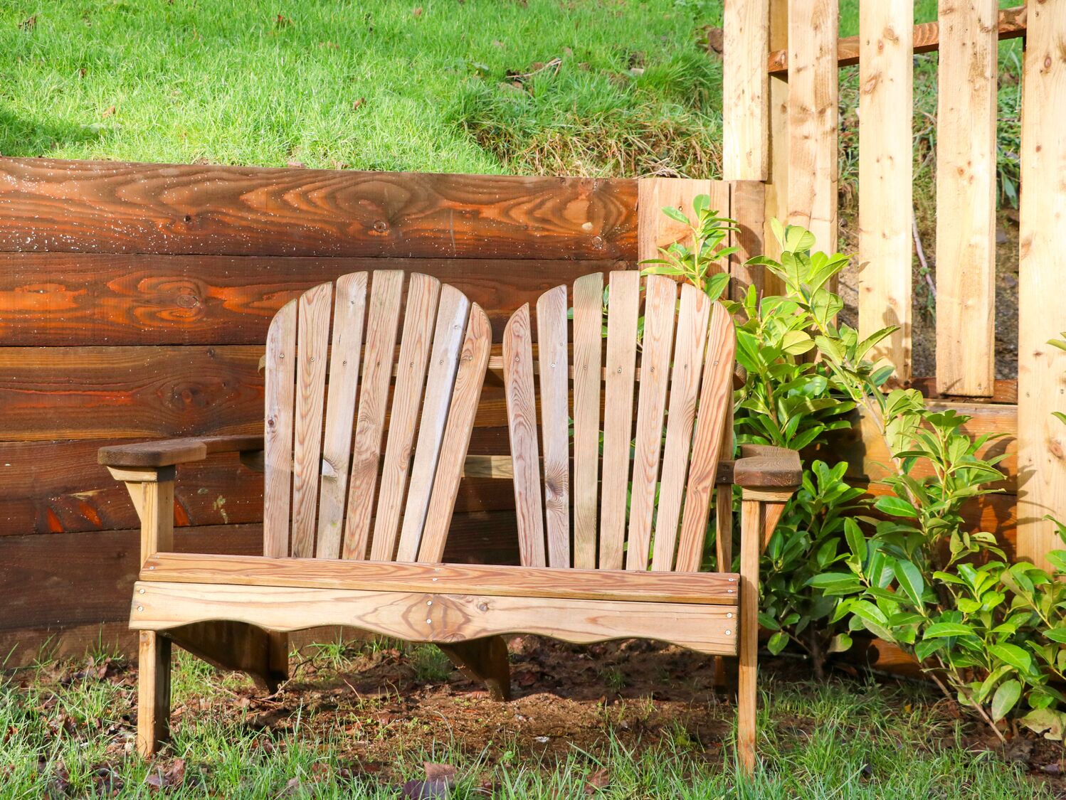 A wooden bench with a curved backrest and armrests, positioned in front of a wooden fence and a green bush.