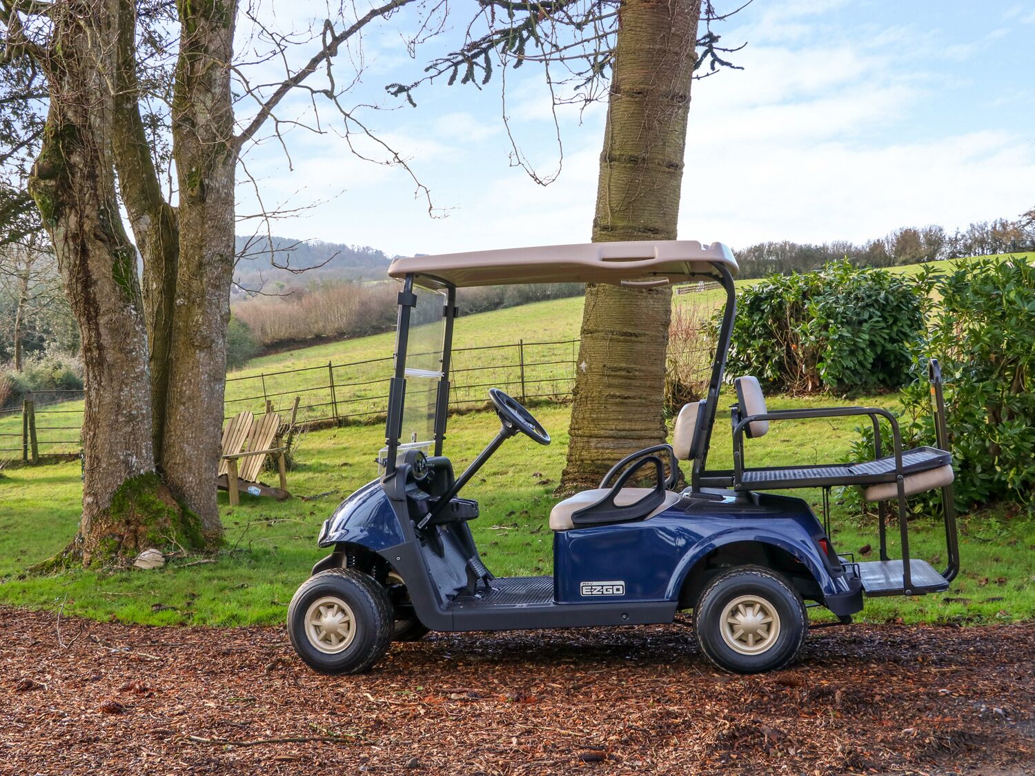 A golf cart parked on a gravel path with trees and a fence in the background.