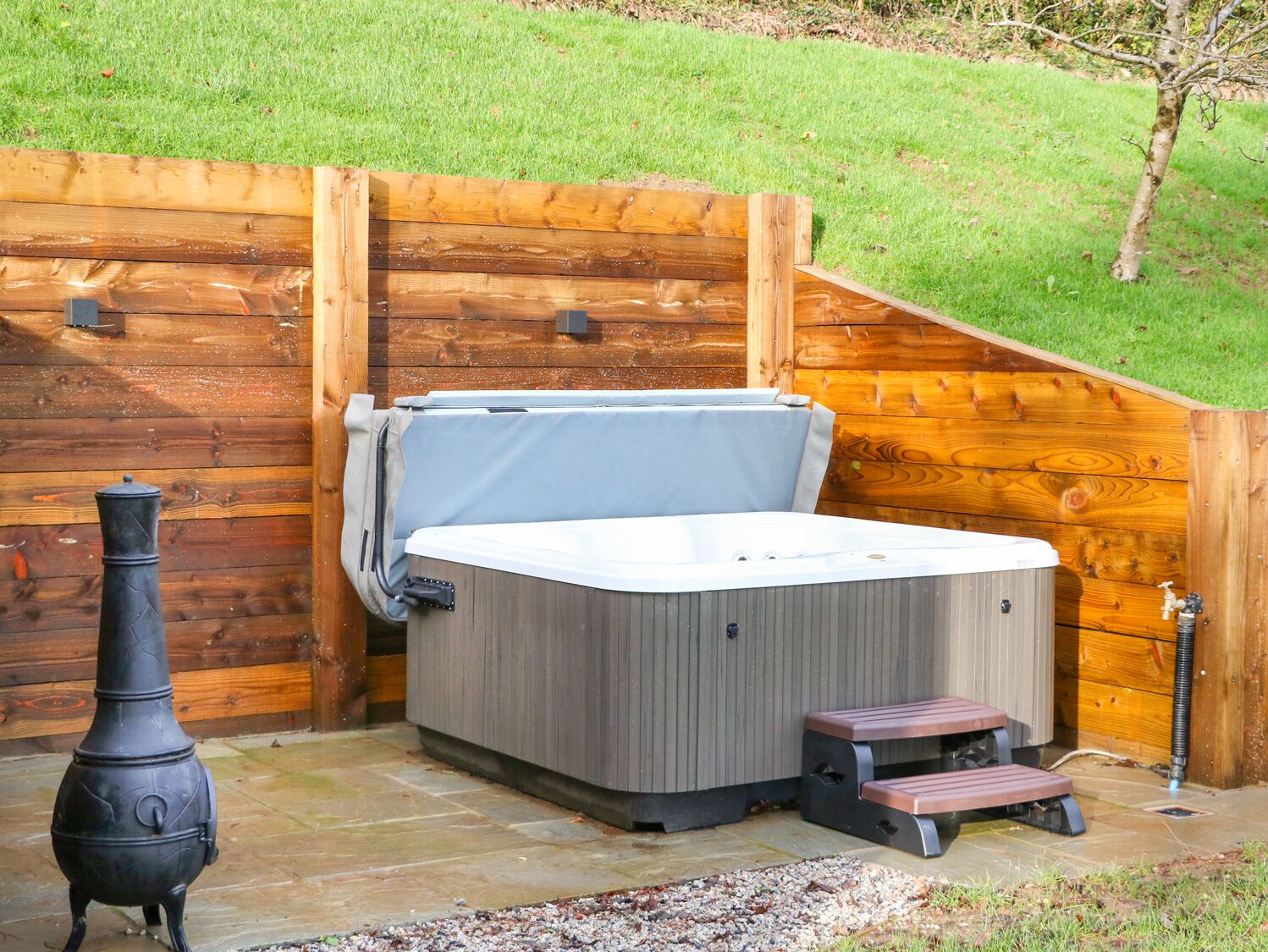 A hot tub in a wooden outdoor setting with a stone floor, a small wooden bench, and a black stove.