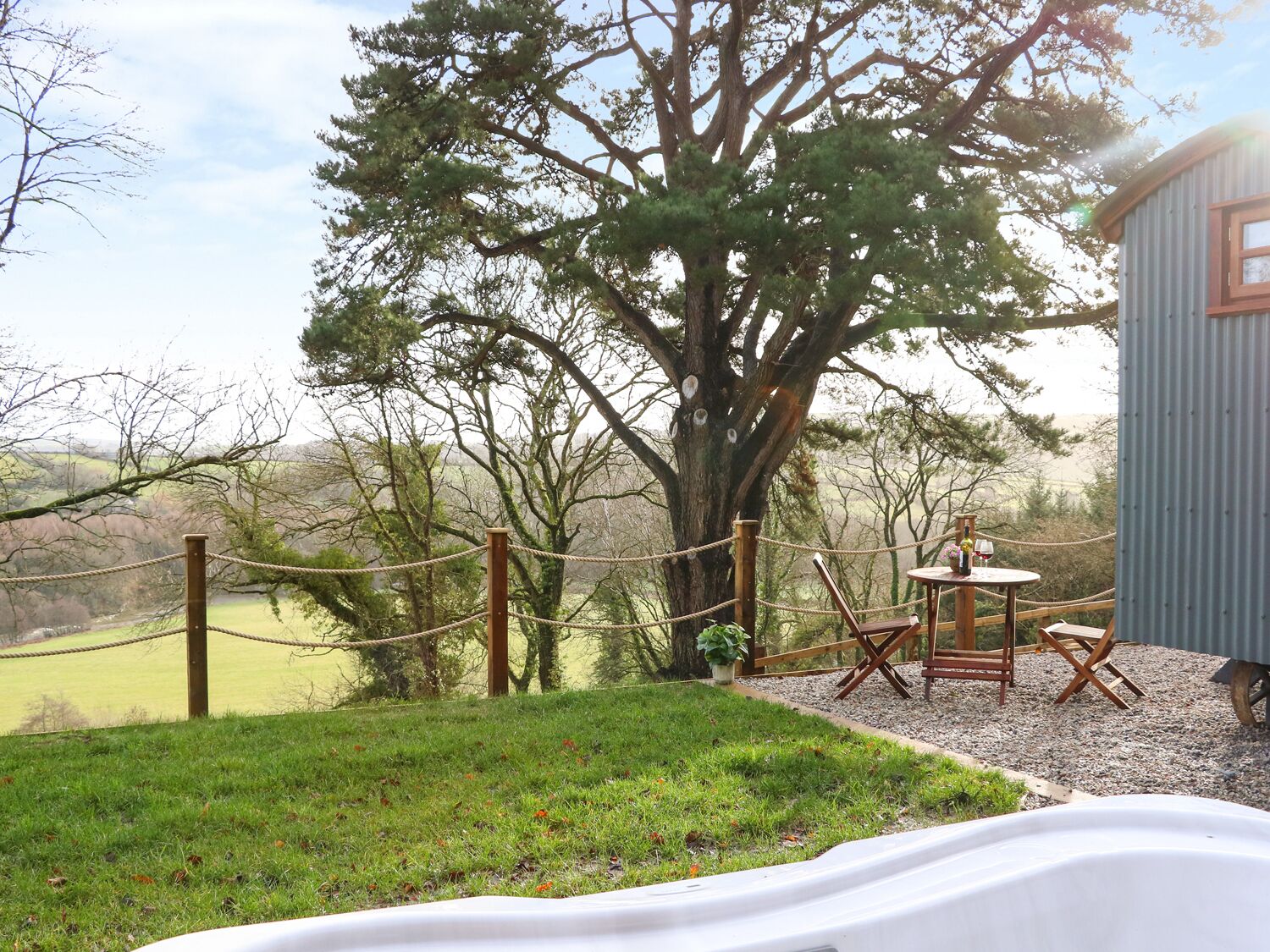 A rustic outdoor seating area with a table and chairs, a large tree, and a view of the countryside.