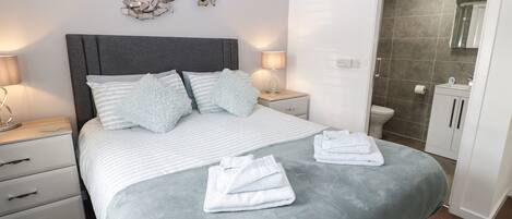 Un dormitorio moderno con una cama tapizada en gris, ropa de cama a rayas blancas y almohadas mullidas. Hay dos mesitas de noche con lámparas, una lámpara de pared y un baño visible a través de una puerta abierta.