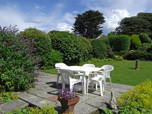 Un patio jardín con mesa y sillas blancas, rodeado de exuberante follaje y plantas florecientes.