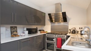 A modern kitchen with grey cabinets, a stainless steel range hood, a gas stove, two ovens, a microwave, and a sink with a kettle and teapot. - Hirfron (Caernarfon)