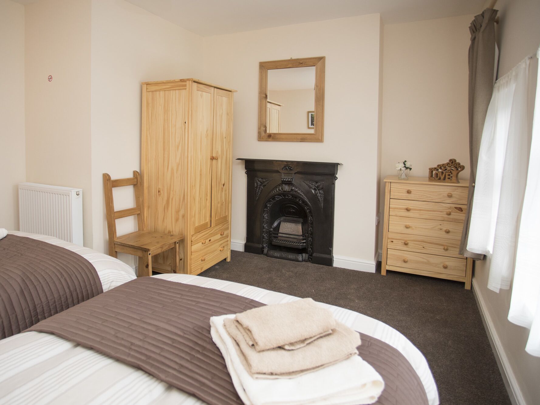 A bedroom with a bed, a wooden wardrobe, a dresser, a fireplace, and a mirror.