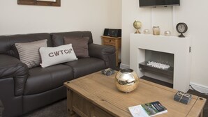 A living room with a dark leather sofa, a wooden coffee table, a flat-screen TV mounted on the wall, a small wooden side table, and a fireplace with decorative items. - Hirfron (Caernarfon)
