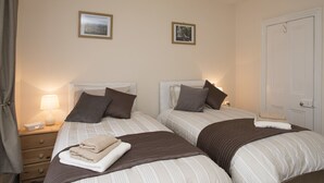 A hotel room with two beds, each with a headboard, pillows, and a blanket. There are two bedside tables with lamps and a small box on one. The walls have two framed pictures. - Hirfron (Caernarfon)