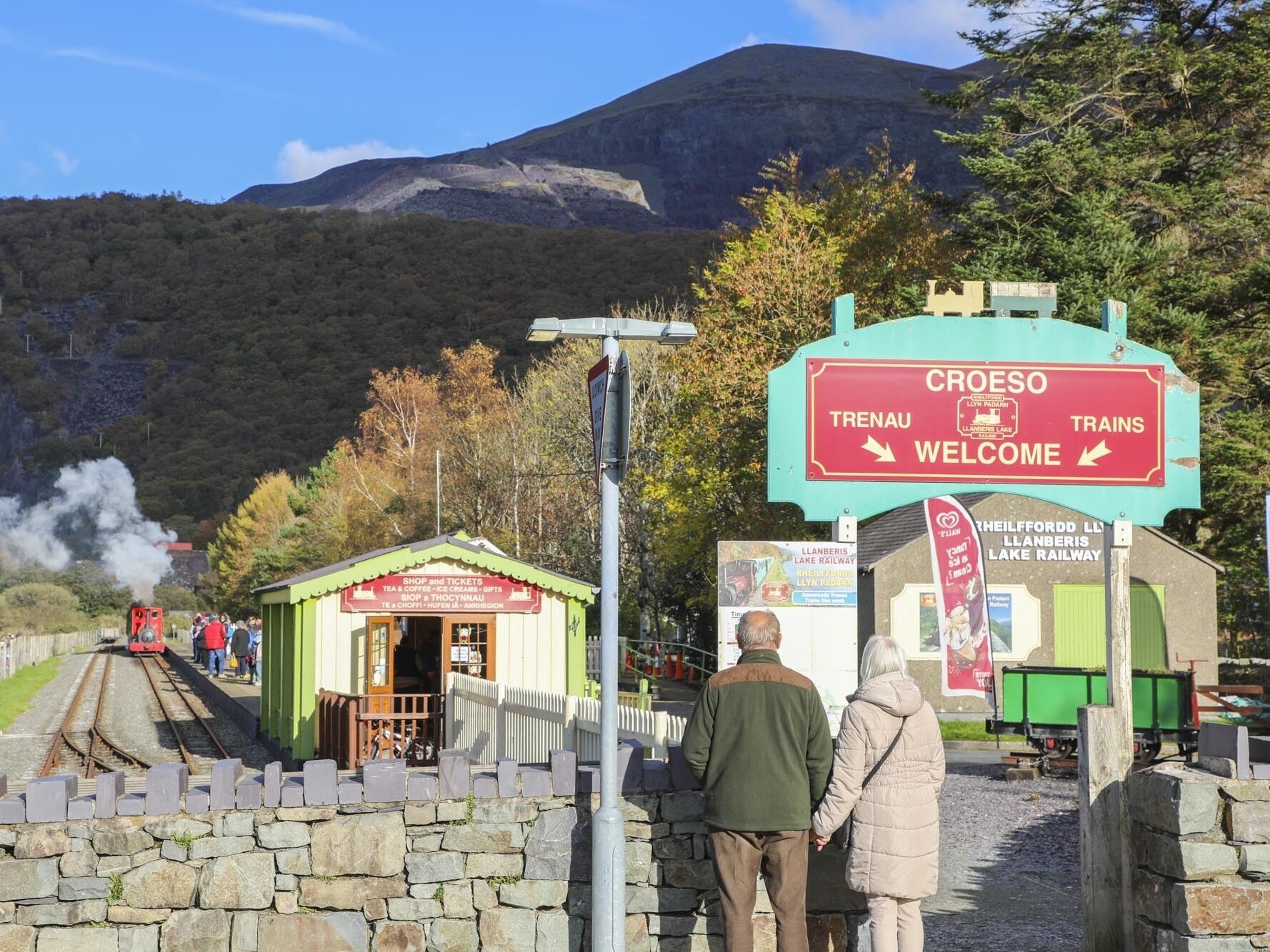 Una estación de tren con un letrero rojo que indica 'CROESO Trenau Welcome' y un letrero verde para 'Llanberis Lake Railway'.