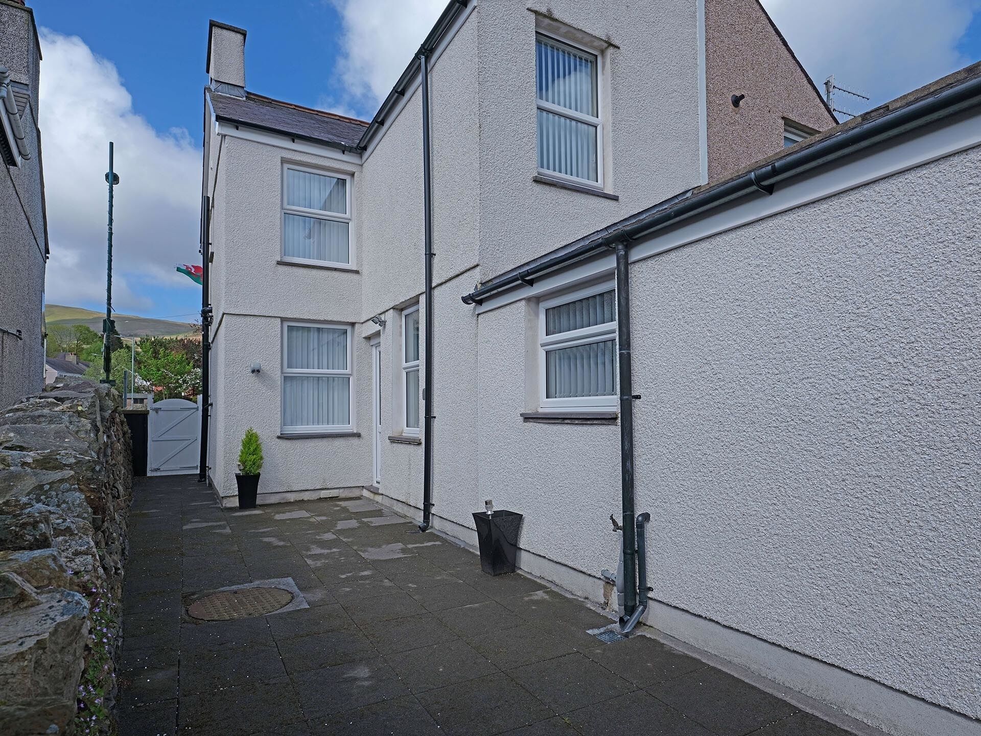 A narrow alleyway between two white buildings with a stone wall on one side and a black gutter on the other.