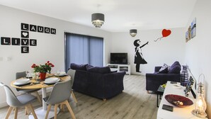 A modern living room with a dining area, featuring a TV, dark sofas, a chandelier, and a wall decal with a girl holding a heart.