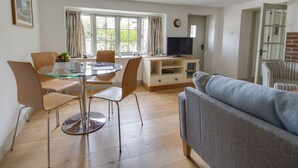 A modern living room with a glass dining table, two chairs, a grey sofa, and a television on a wooden stand. - Little Forge (Wareham)