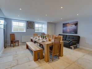A dining area with a wooden table and benches, a black sofa, and a large window with blinds. - Low Tide (Weymouth)
