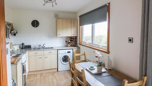 Une cuisine compacte équipée d’un lave-linge, de meubles en bois, d’une table à manger avec des chaises et d’une grande fenêtre qui laisse entrer la lumière naturelle.