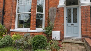 Ein rotes Backhaus mit einer weißen Tür, die mit der Nummer 8 gekennzeichnet ist, ein großes Fenster mit weißen Rahmen und ein kleiner Garten mit verschiedenen Pflanzen und Blumen.