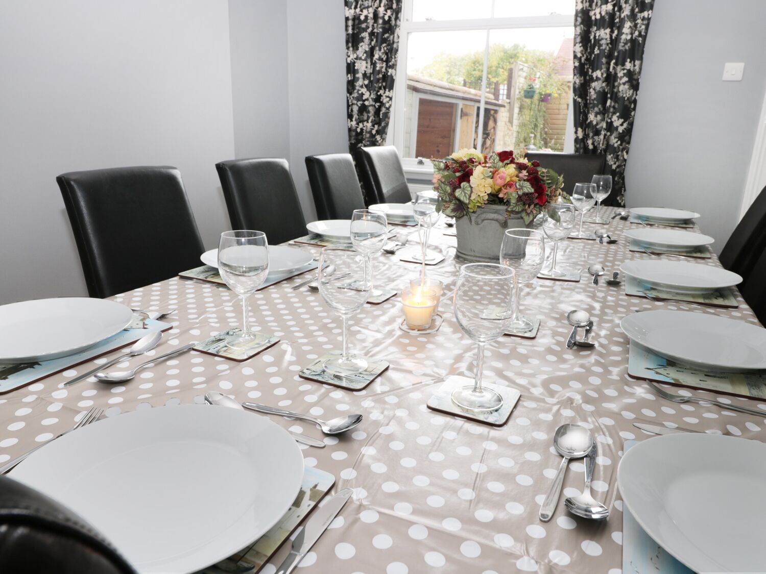 A dining table set for six with white plates, cutlery, and glasses, a candle centerpiece, and a window with curtains.