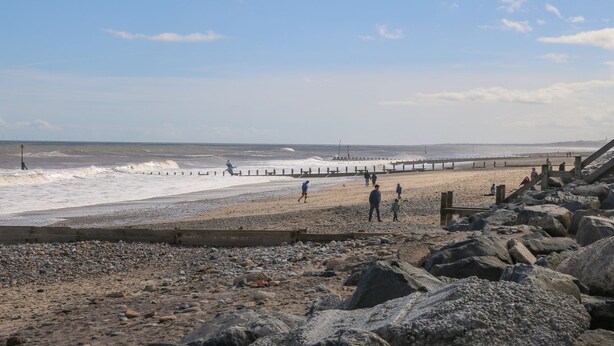 Ein felsiger Strand mit einer Holzpromenade, die zum Meer führt, ein Leuchtturm in der Ferne und ein klarer Himmel.