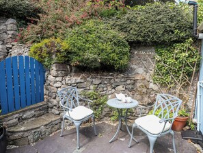 A stone wall with a blue gate, two metal chairs with cushions, a small round table with a teapot and cup, and a potted plant.