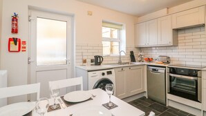 A modern kitchen with white cabinetry, a built-in oven, and a dining area set for four with white tableware and glasses. - Maisy Cottage (Weymouth)