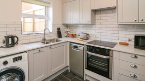 A compact kitchen with white cabinets, a built-in washing machine, a dishwasher, a toaster, and a window with a view of the outside. - Maisy Cottage (Weymouth)