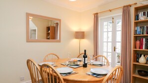A dining room with a wooden table set for six, a mirror on the wall, a bookshelf with books, and a lamp. - Louisa Cottage (Weymouth)