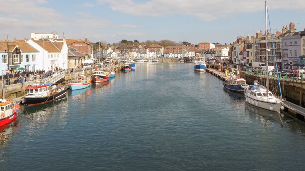 A harbor with boats docked along the waterfront, buildings lining the shore, and a clear sky. - Louisa Cottage (Weymouth)