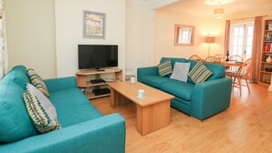 A living room with teal sofas, a wooden coffee table, a flat-screen TV, and a dining area with a wooden table and chairs. - Louisa Cottage (Weymouth)