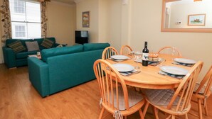 A dining area with a wooden table set for four, surrounded by wooden chairs. A television is mounted on the wall in the background. - Louisa Cottage (Weymouth)