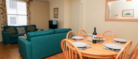 A dining area with a wooden table set for four, surrounded by wooden chairs. A television is mounted on the wall in the background.