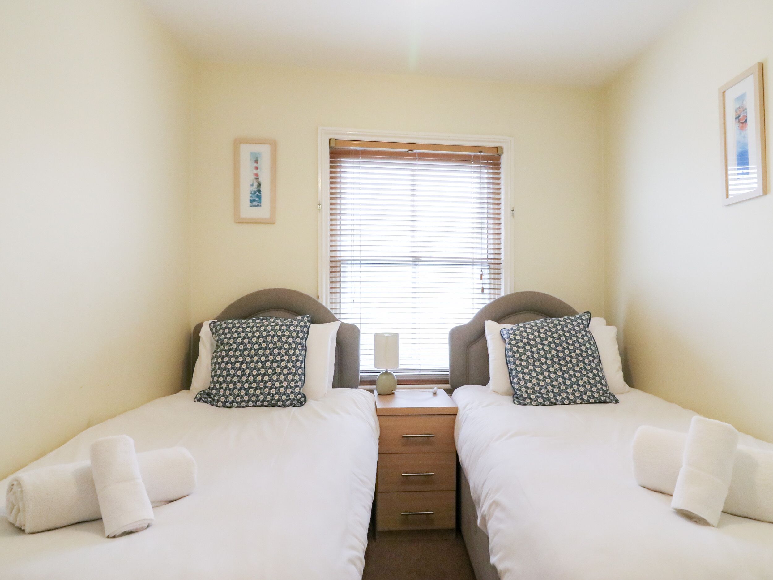 A twin room with two single beds, a small bedside table, a window with blinds, and a framed picture on the wall.