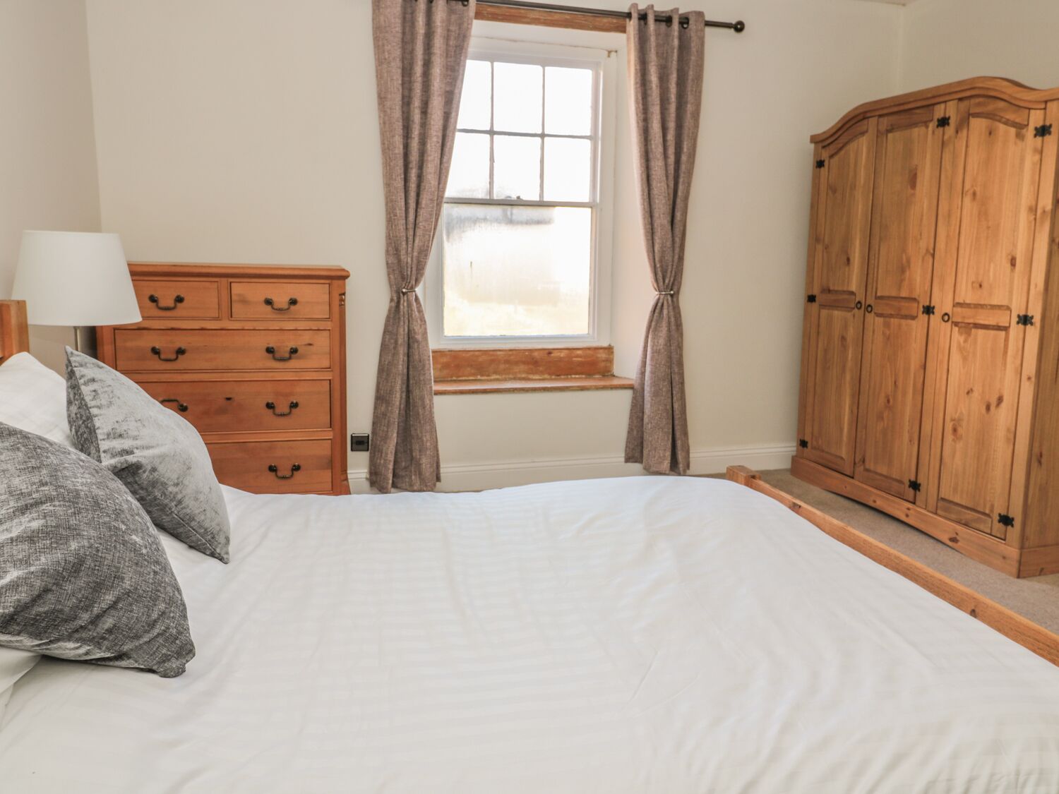 A bedroom with a bed, a wooden dresser, a window with curtains, and a wooden wardrobe.
