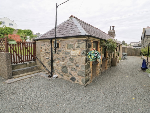 A stone building with a sloped roof, a gravel driveway, a wooden gate, and a hanging flower basket. - Fir Tree Cottage (Criccieth)