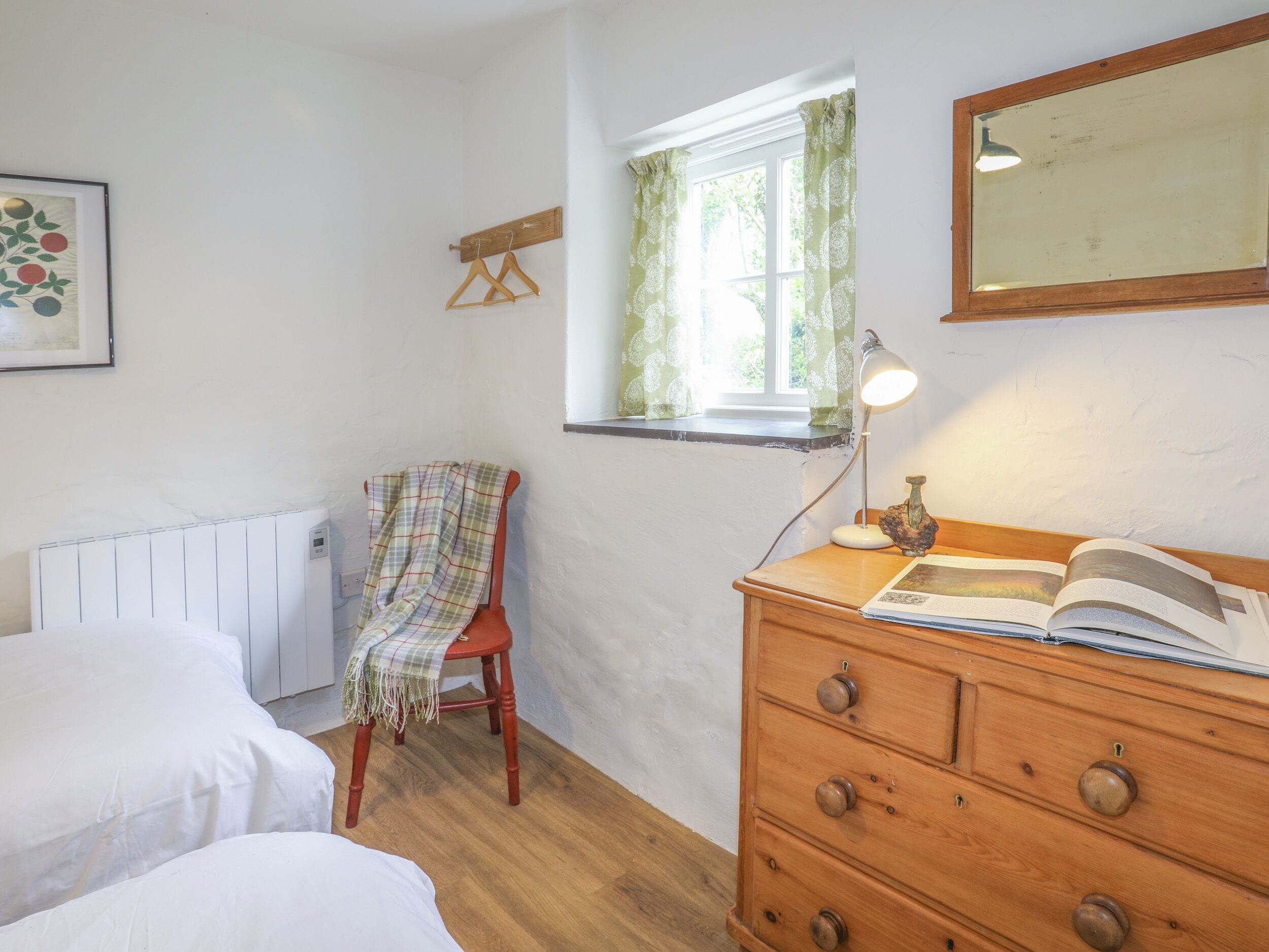 A bedroom with a bed, a wooden dresser, a chair, a radiator, a window with curtains, and a mirror on the wall.