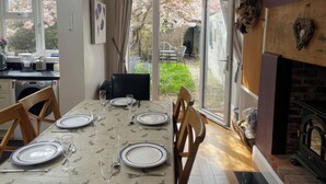 En matplats med ett bord för fyra personer, en öppen spis och utsikt över en trädgård genom glasdörrar.