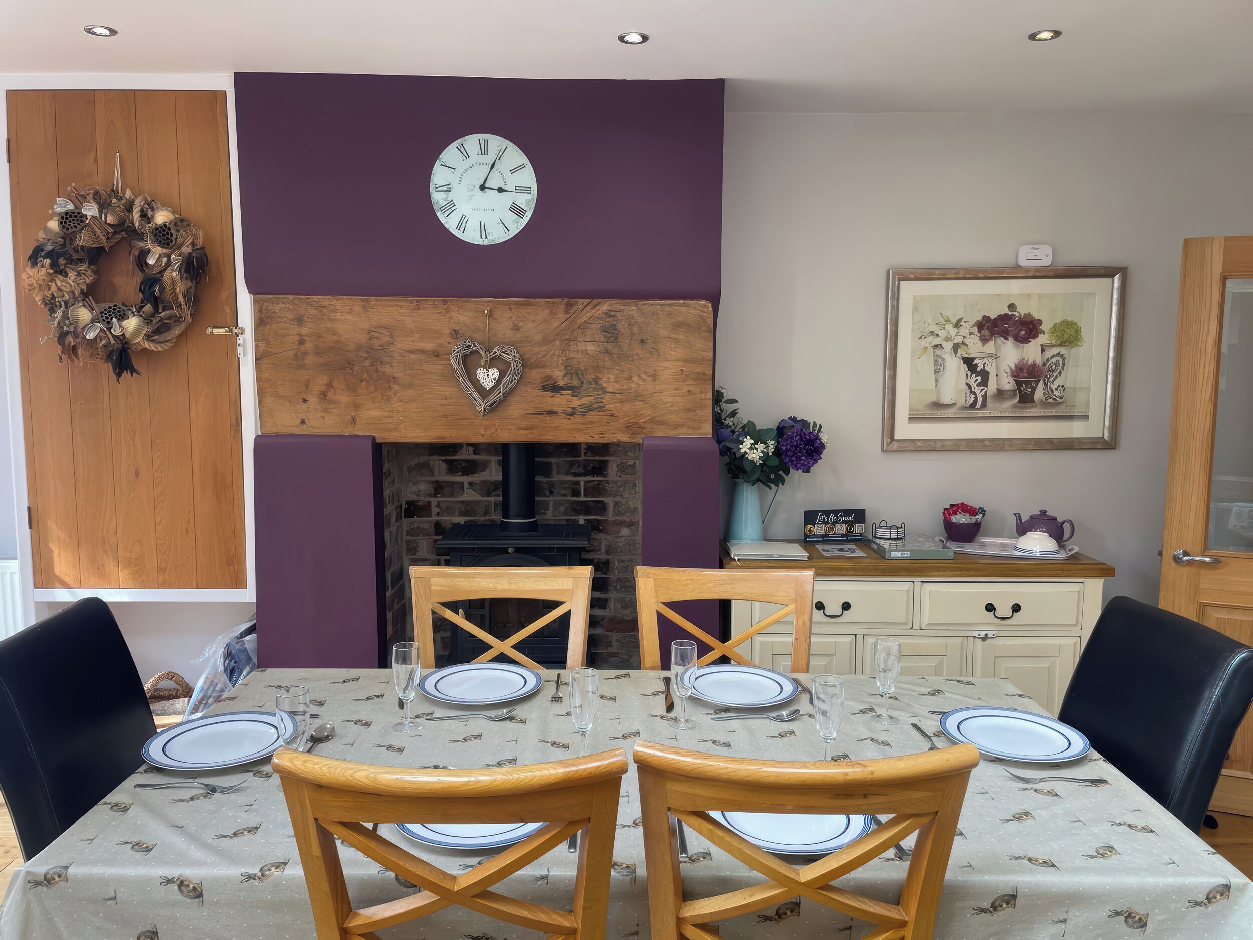 A dining room with a wooden table set for four, a clock above the fireplace, and a decorative wreath on the wall.