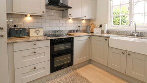 A modern kitchen with white cabinets, a built-in oven, a countertop with a kettle, and a window with a view of greenery. - Sunlight Soap Cottage (Wirral)
