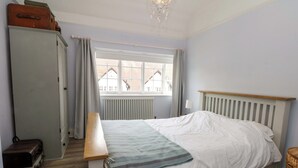 A bedroom with a bed, a dresser, a window with curtains, a radiator, and a chandelier. - Sunlight Soap Cottage (Wirral)