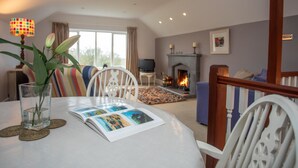A cozy living room with a fireplace, a dining table with a magazine, and a colorful lamp. - Maes y Cregyn (Marianglas)