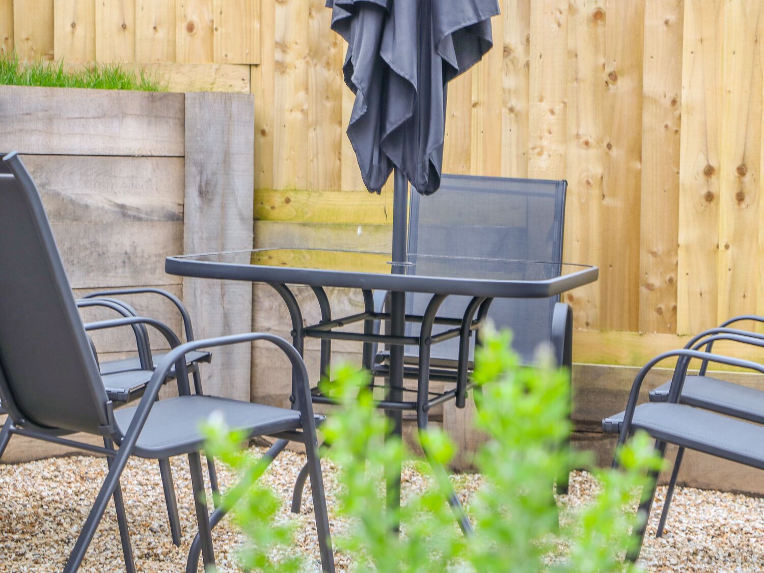 A patio area with a table and four chairs, a wooden fence in the background, and a grassy area above the fence.