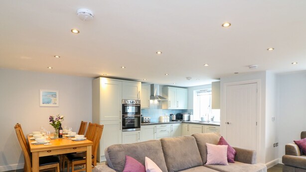 A modern living room with a grey sofa, a dining table with chairs, and a kitchen area in the background. - Exmoor View (South Molton)