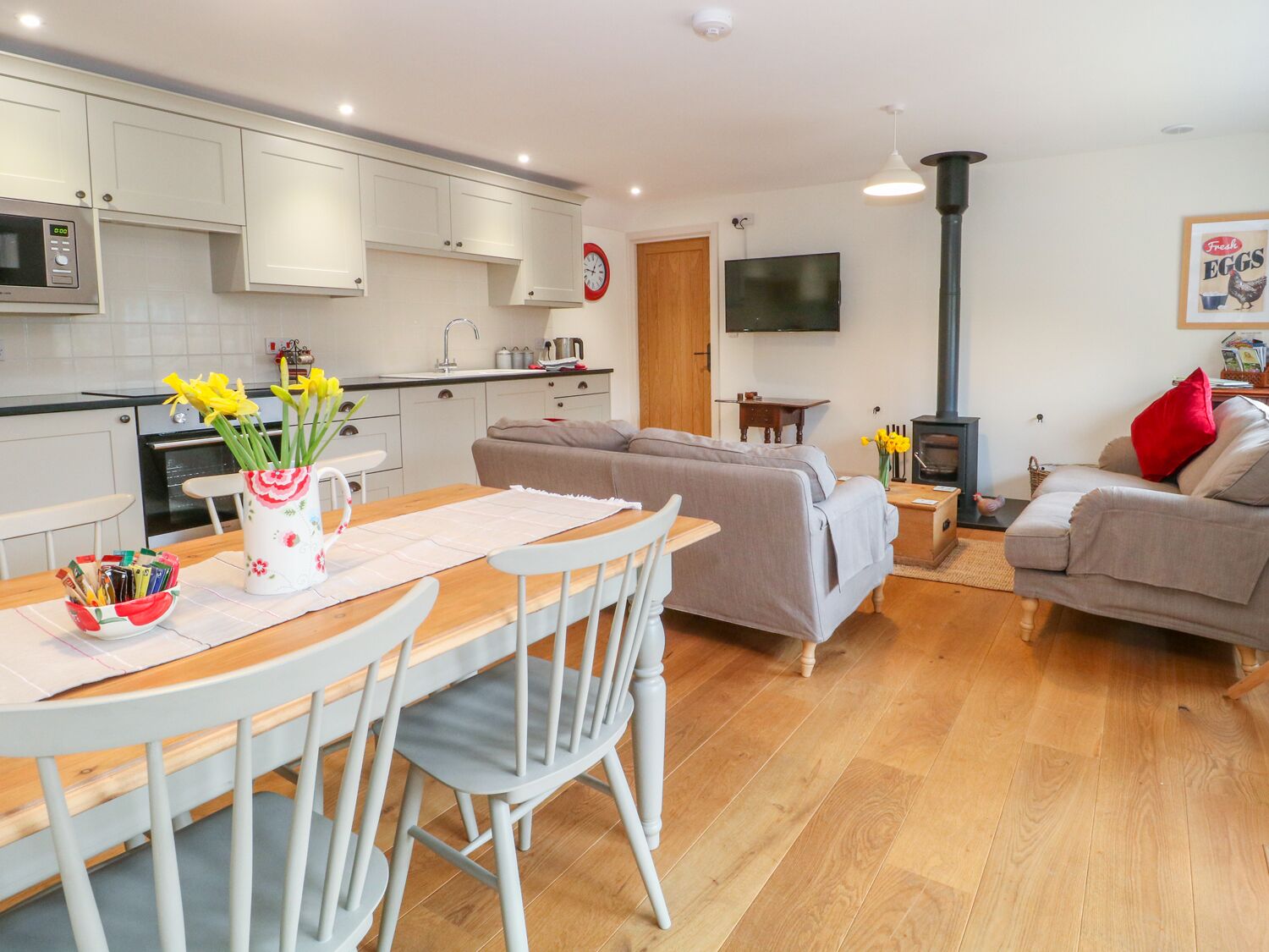 A modern kitchen with white cabinets, a dining table with chairs, a sofa, a TV, and a fireplace.