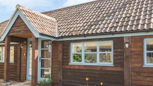 Une maison en bois avec un toit en tuiles, une porte d’entrée et des fenêtres. Devant la maison, on trouve des parterres de fleurs avec des plantes et des fleurs.