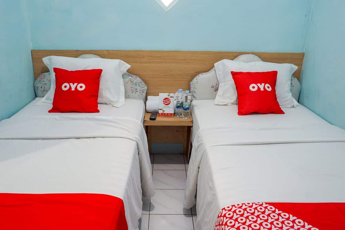 Une chambre d’hôtel avec deux lits, chacun agrémenté d’un oreiller rouge portant l’inscription « OYO » et recouvert d’une couverture blanche. Une table de chevet se trouve entre les deux lits, sur laquelle reposent une télécommande, une bouteille d’eau et un panneau indiquant : « À QUOI PENSEZ-VOUS ? ».