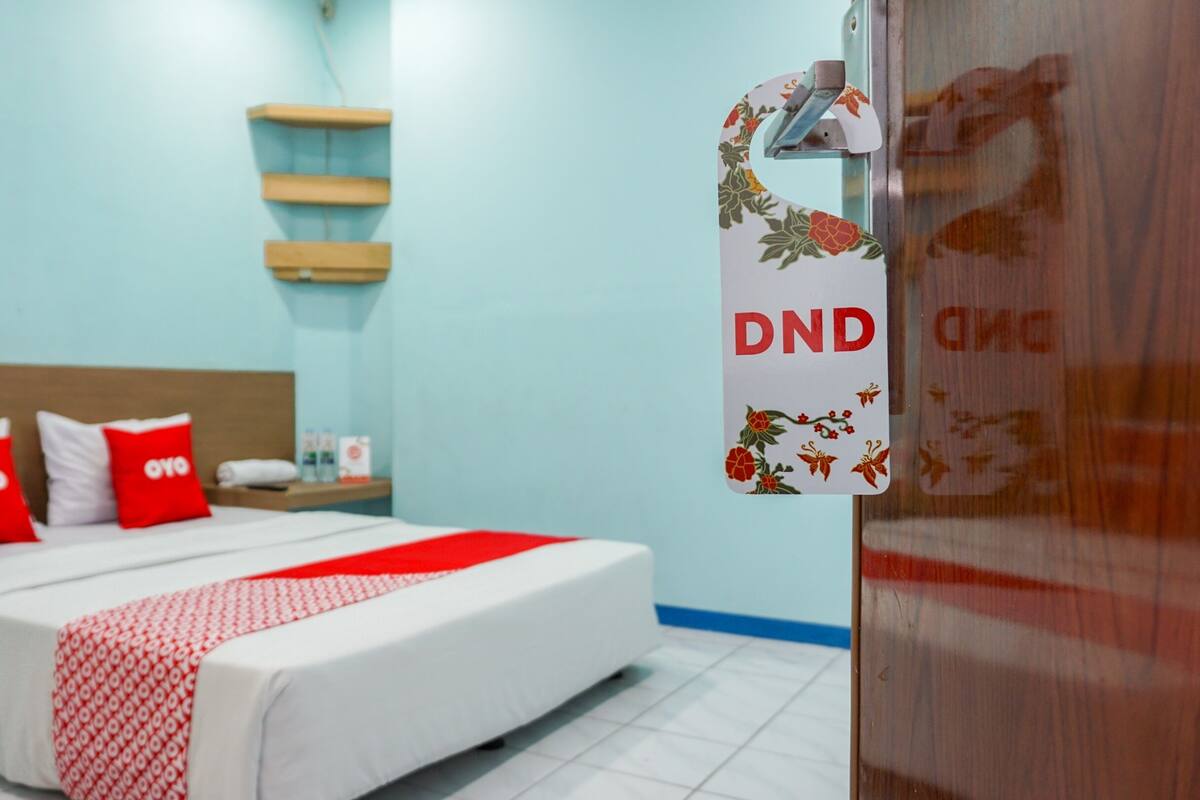 Une chambre d’hôtel avec un lit, deux oreillers rouges, une tête de lit en bois, une table de chevet et une étagère fixée au mur.