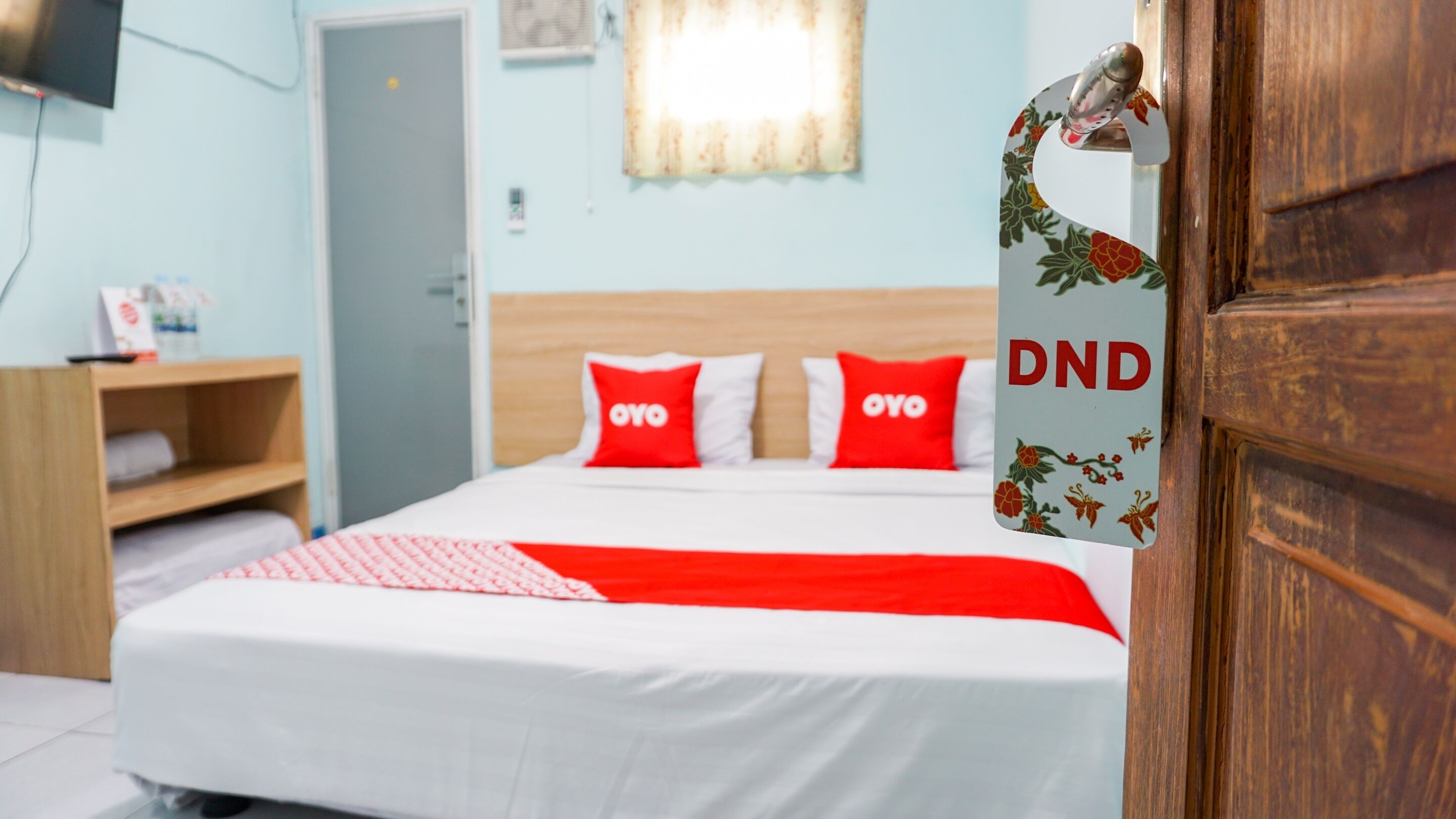 A hotel room with a bed, two red pillows with 'OYO' branding, a wooden headboard, a wooden door with 'DND' on it, and a television on the wall.
