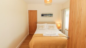 A bedroom with a wooden door, a bed with white bedding, a yellow throw blanket, a bedside lamp, and a painting on the wall. - Llain Bach (Llangefni)